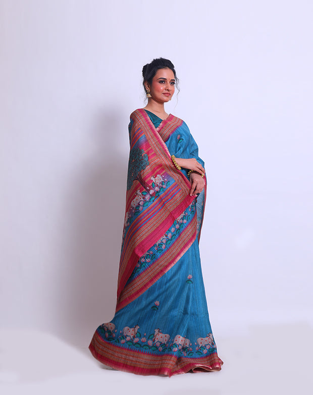 The blue semi-Tussar saree with a pink border featuring Pichwai design adds a touch of cultural richness and elegance to the ensemble - BLN00855