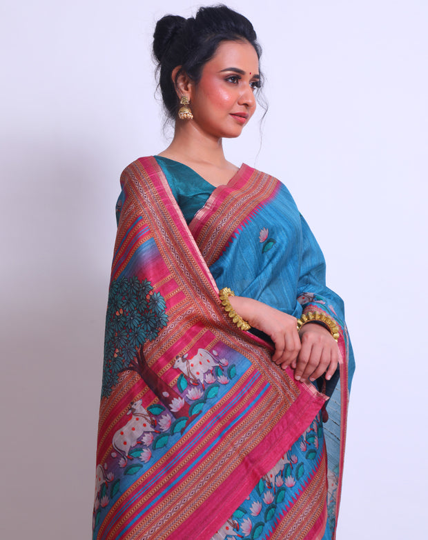 The blue semi-Tussar saree with a pink border featuring Pichwai design adds a touch of cultural richness and elegance to the ensemble - BLN00855